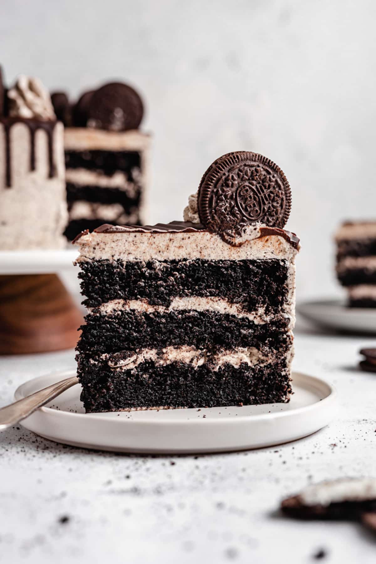Slice of a triple layer chocolate Oreo cake on a white plate with the full cake in the background.