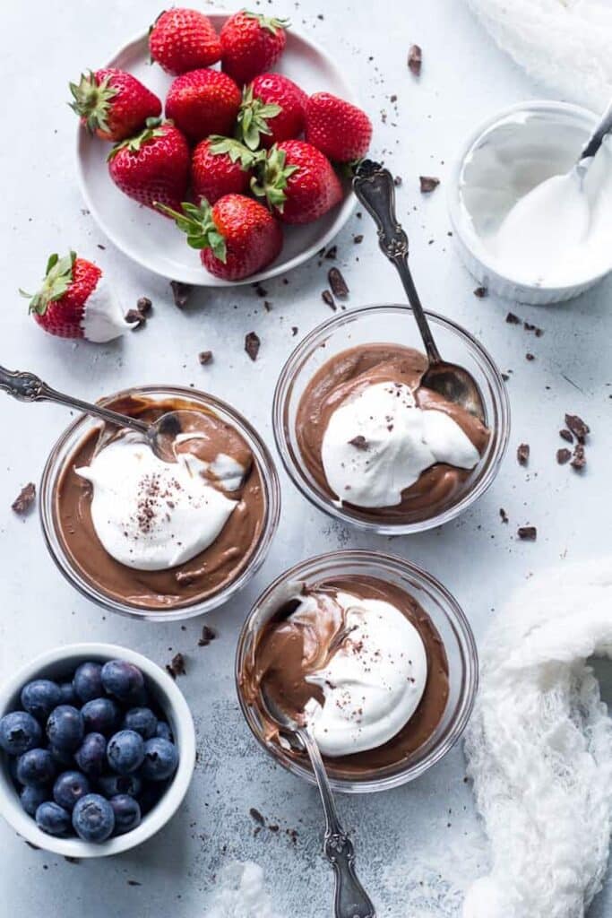 Three chocolate puddings in glass cups with spoons sticking out and berries in bowls. 