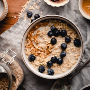 Creamy oatmeal topped with peanut butter and blueberries with it's ingredients in bowls surrounding it.