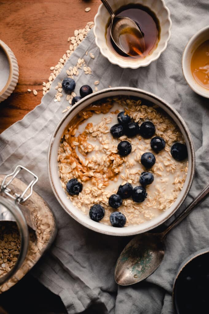 Creamy oatmeal topped with peanut butter and blueberries with it's ingredients in bowls surrounding it.