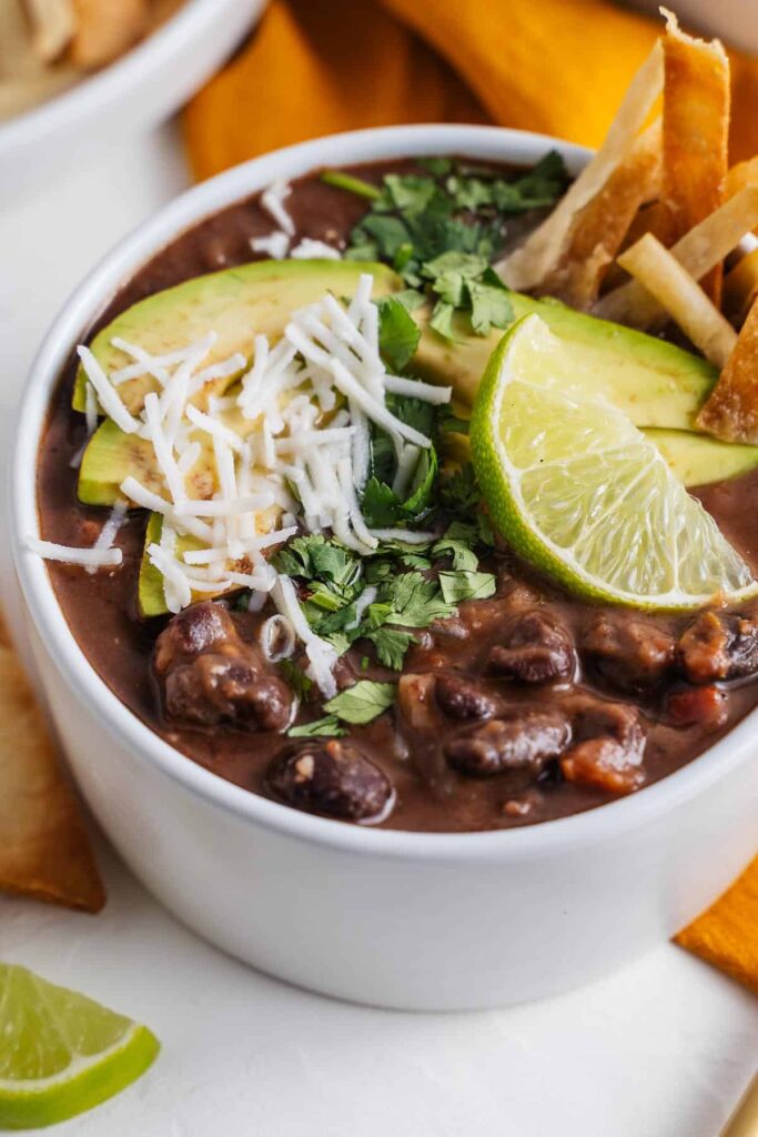 A bowl of black bean vegan soup garnished with avocado slices, shredded cheese, cilantro, a lime wedge, and served with tortilla strips.
