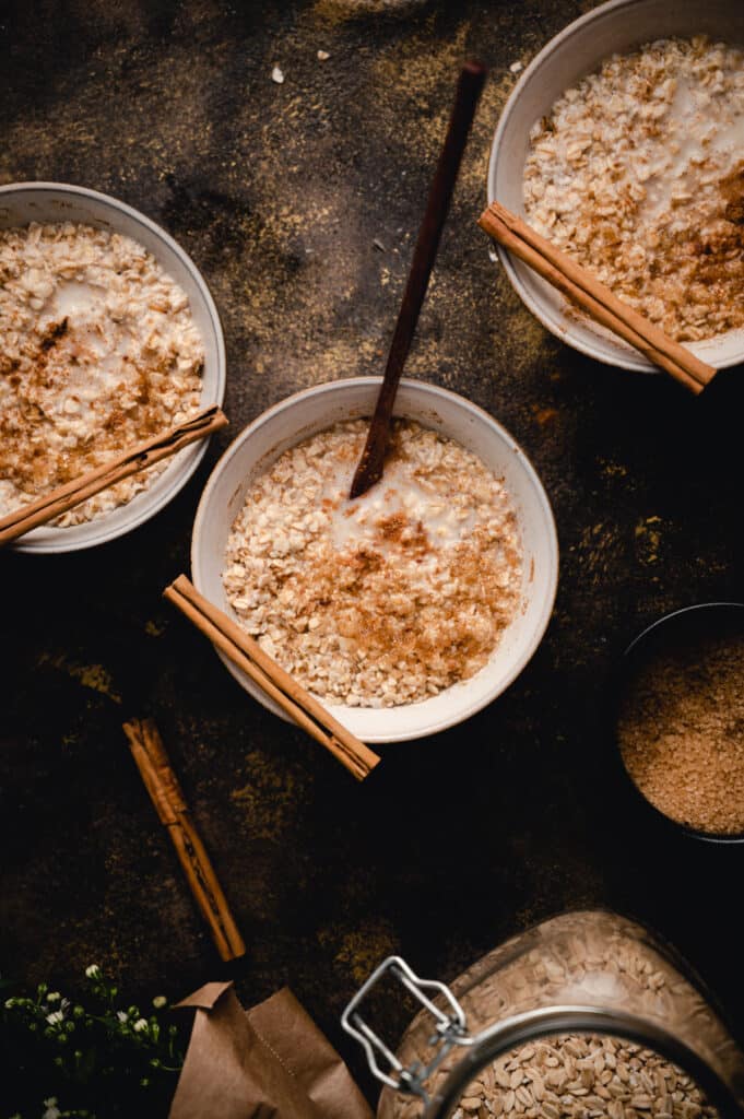 Three bowls of brown sugar oatmeal with cinnamon sticks on top and a wooden spoon sticking out.