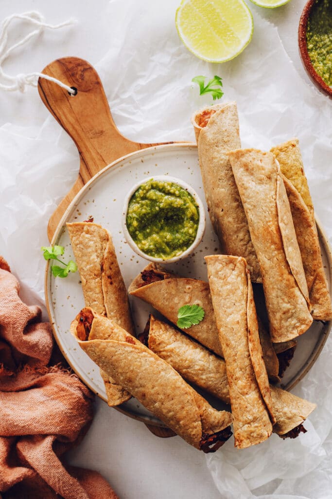 Wooden platter covered in crispy taquitos with a bowl of guacamole.