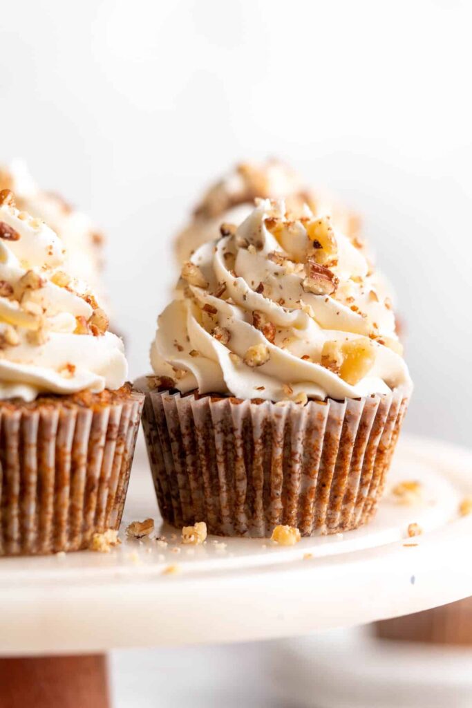 Two carrot cupcakes with inches of swirled cream cheese frosting and chopped walnuts sprinkled on top.