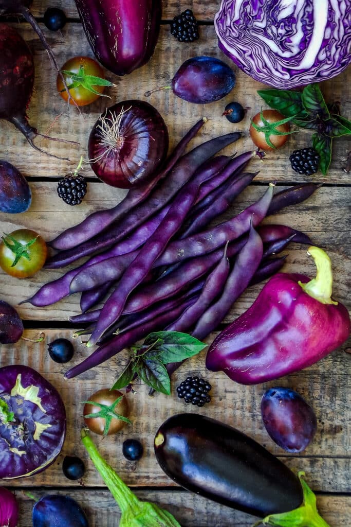 Wooden table covered in purple veggies and fruit with purple beans in the middle.