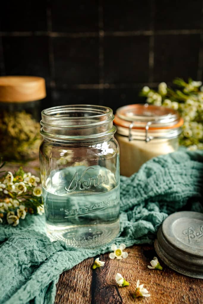 Pint glass jar part filled with water on a wooden table with a teal cloth and flowers.