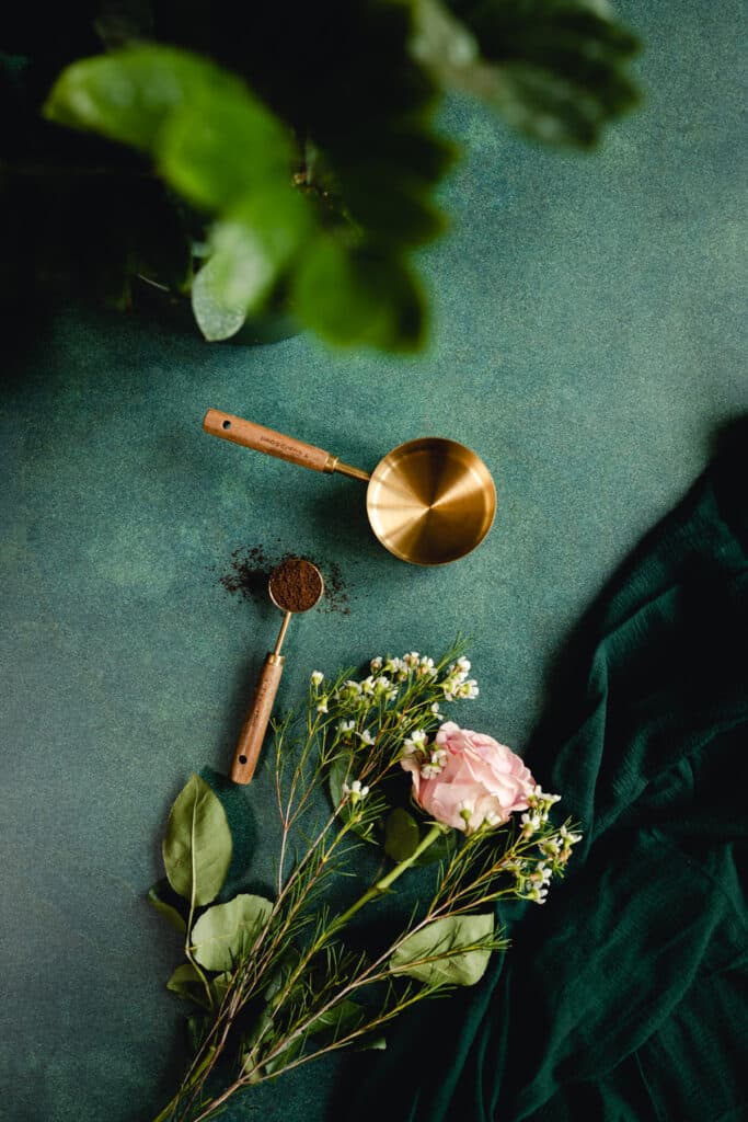 Wood and brass measuring spoon and cup with light pink rose on a green surface.