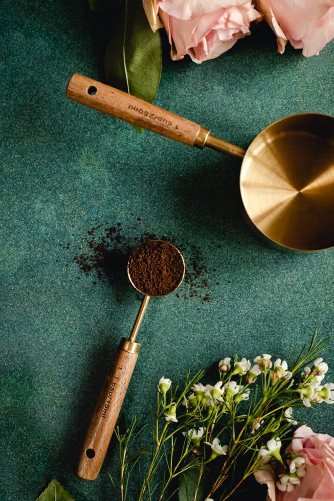One tablespoon filled with coffee on a green surface with flowers laying next to it.