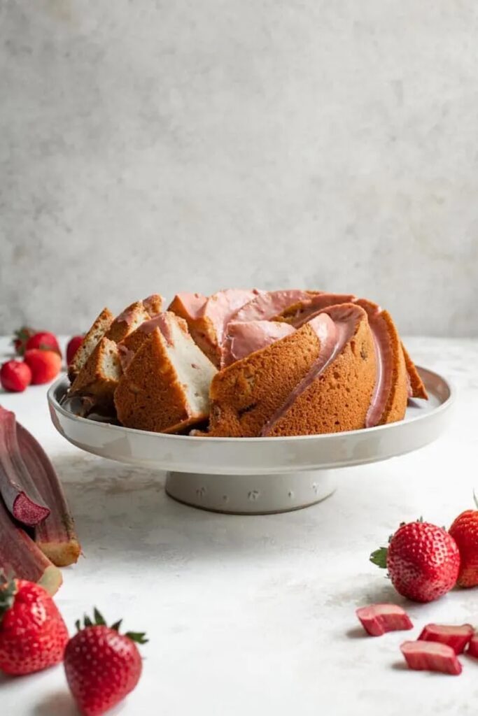 Rhubarb bundt cake on a white cake stand with strawberries surrounding it.