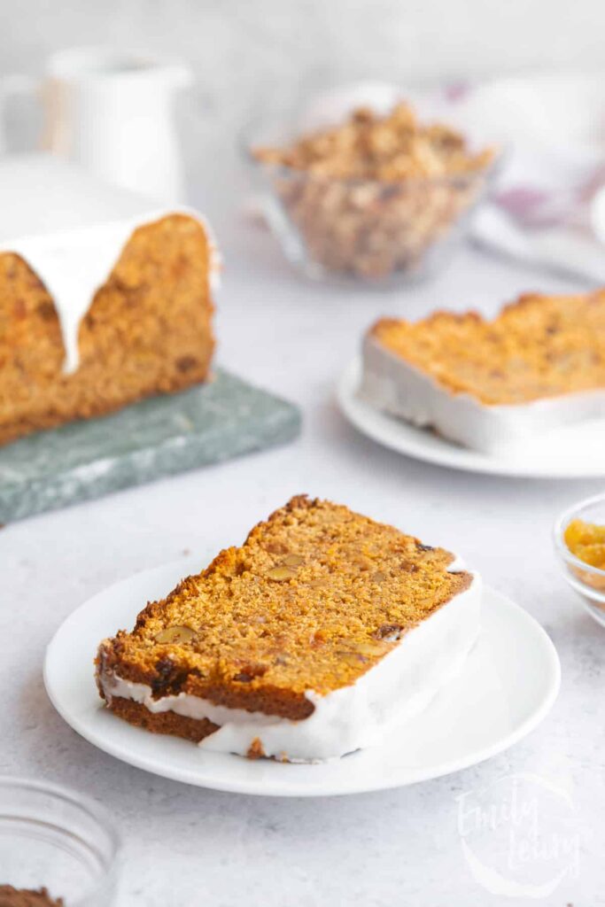 A slice of vegan carrot loaf cake on a white plate with the remaining cake in the background.