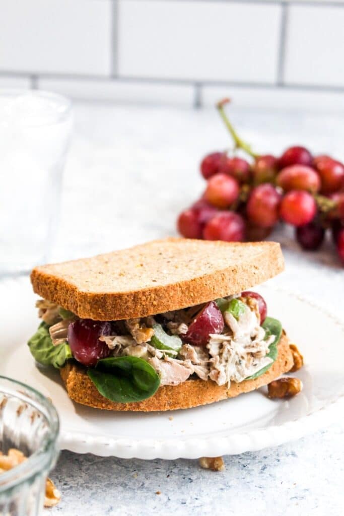 Jackfruit chicken salad sandwich on a white plate with sliced red grapes falling out.