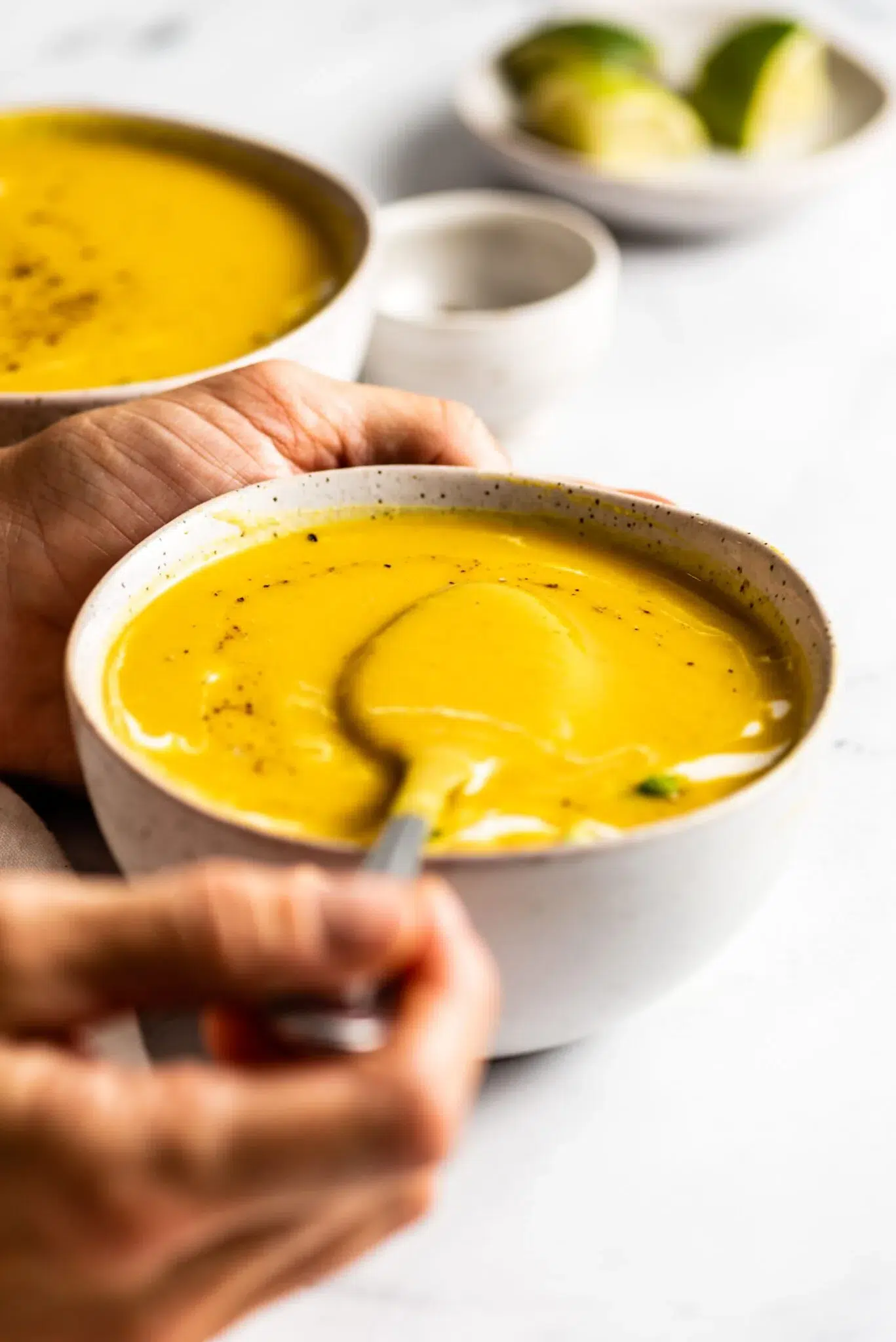 Close-up of a hand holding a bowl of creamy vegan pumpkin soup with a spoon, another bowl, and lime wedges in the background.