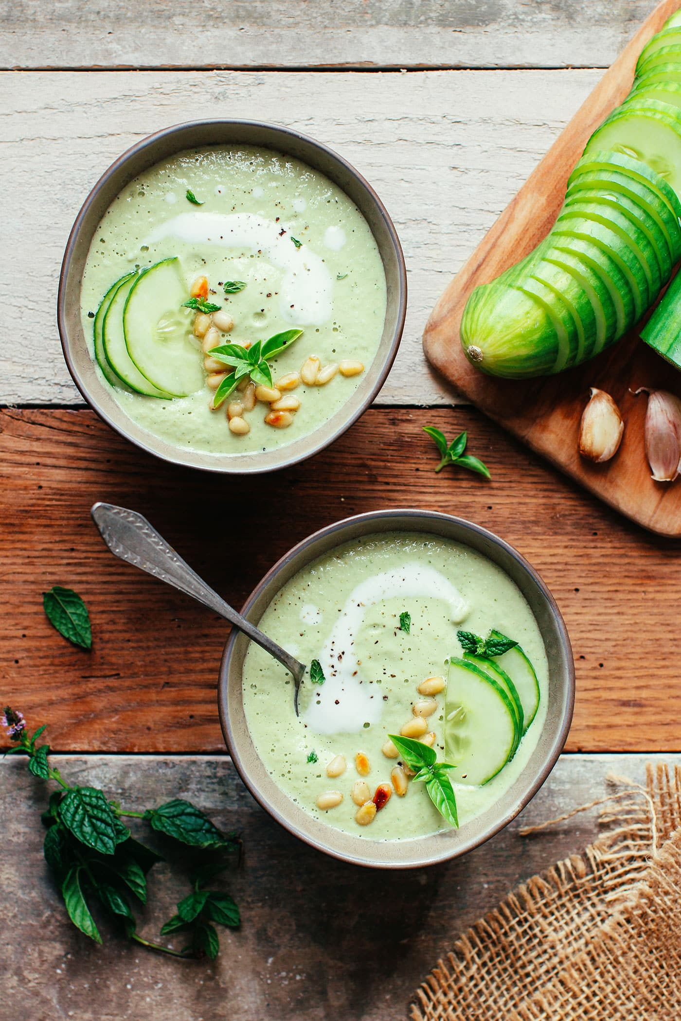 Two bowls of creamy cucumber soup from the best vegan soup recipes, garnished with pine nuts and fresh herbs, with sliced cucumbers and garlic on a wooden board.