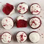 A dozen red velvet cupcakes with white frosting and two with bites taken out.
