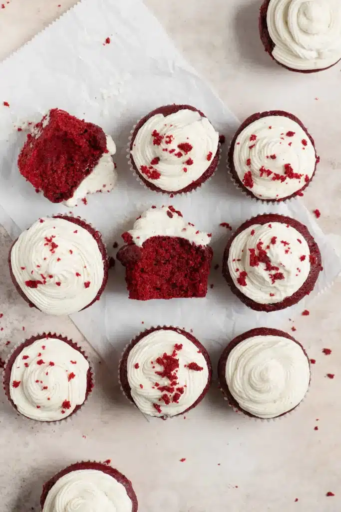 A dozen red velvet cupcakes with white frosting and two with bites taken out. 
