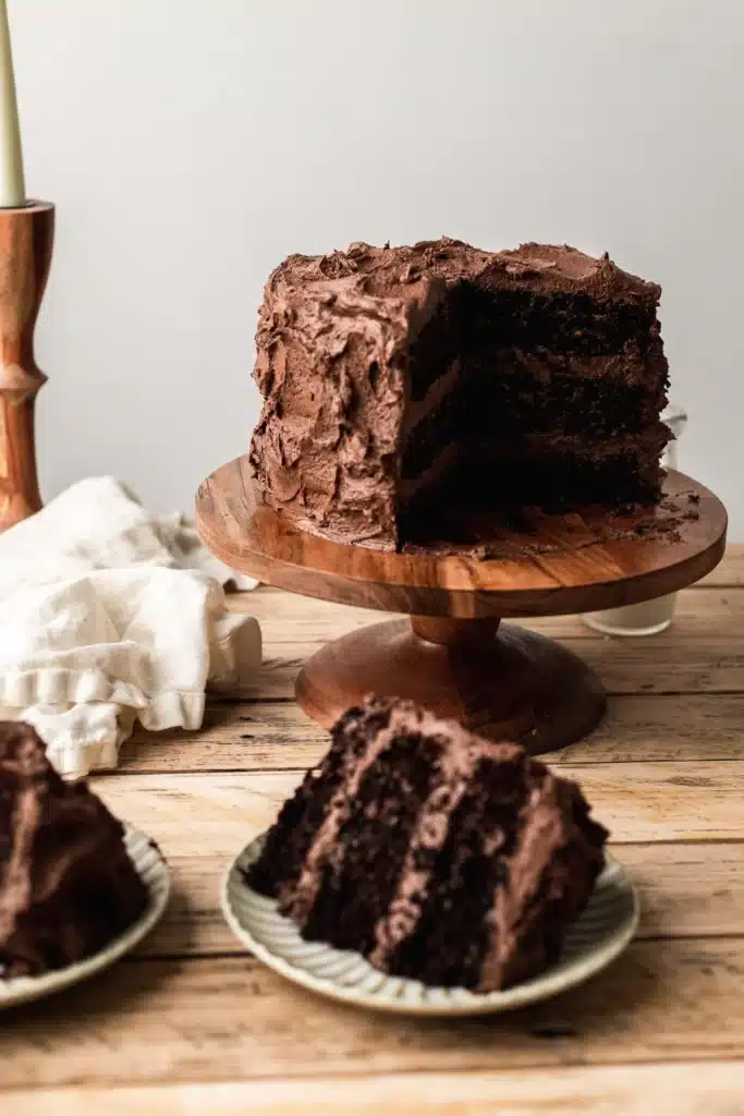 Three layer chocolate cake on a wooden cake stand with two pieces sliced out on plates.