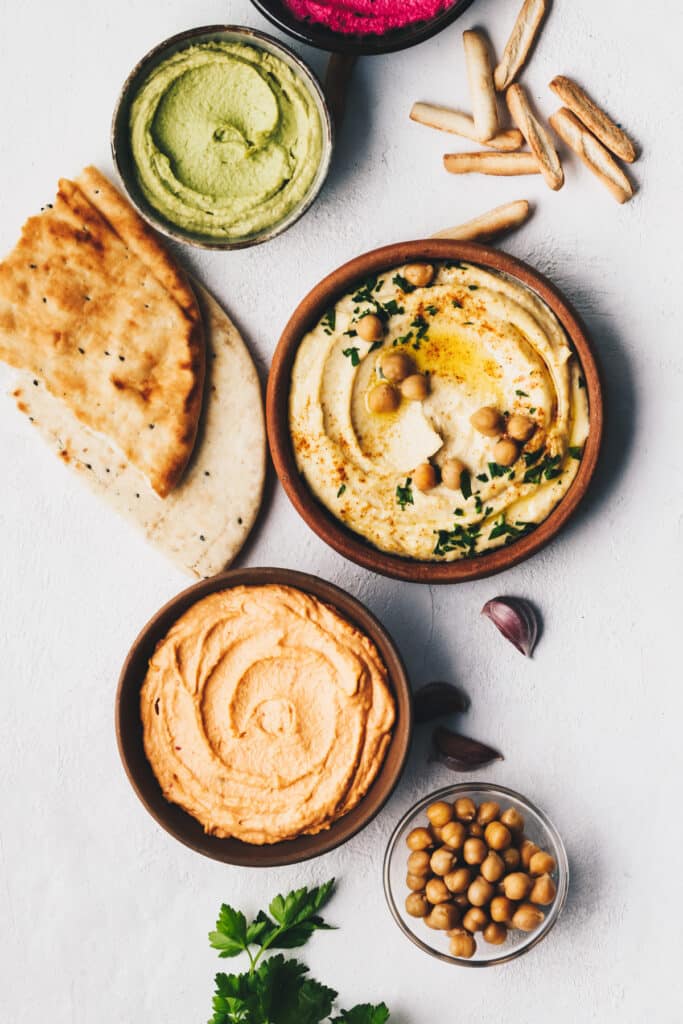 Hummus with drizzled olive oil and chickpeas on top with other types of hummus in smaller bowls.