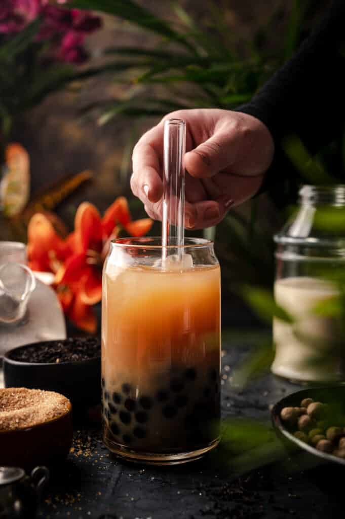 Easy milk tea recipe with bubbles in a clear glass cup surrounded by tropical plants.