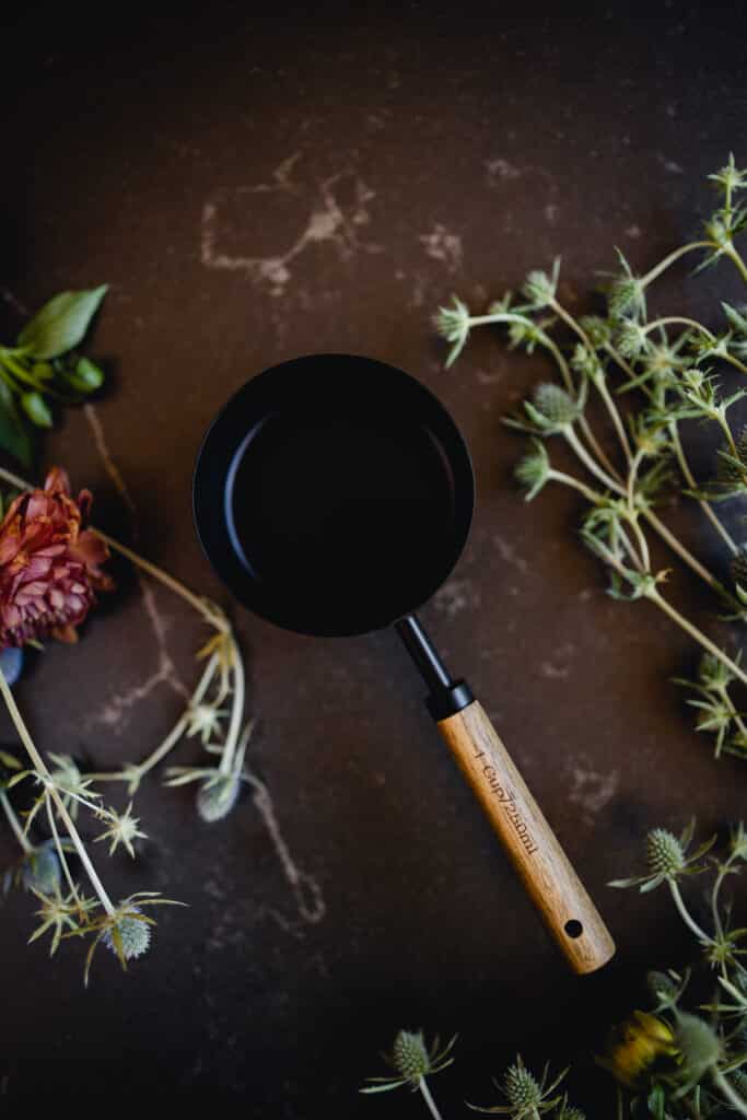 Black measuring cup with wooden handle on soapstone counter with flowers surrounding it.