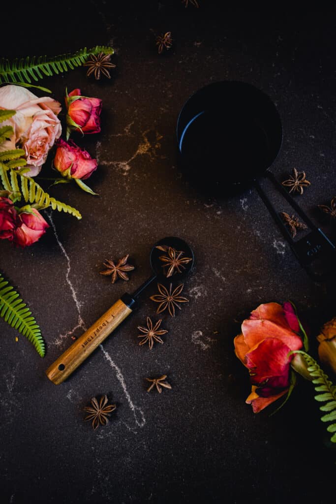 Dark surface with black measuring cup and tablespoon with roses and star anise surrounding them.