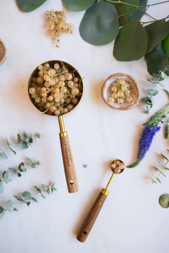 Brass and wood measuring cup and spoon with white currants on a marble counter.