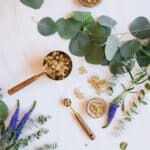 Flatlay of wood and gold teaspoon and cup with eucalyptus and purple flowers.