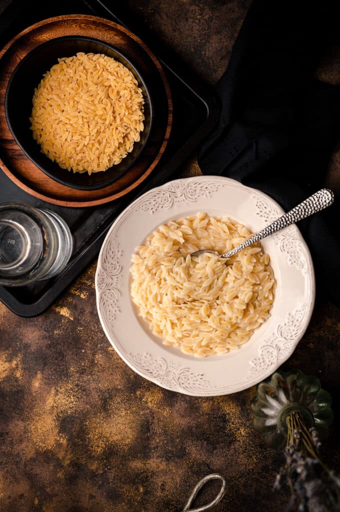 White soup bowl of cooked orzo with a smaller bowl of uncooked orzo.