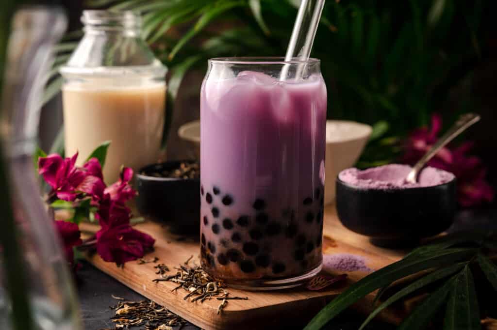 Glass of taro milk tea with boba on a wooden cutting board with a glass straw sticking out.