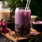Glass of taro milk tea with boba on a wooden cutting board with a glass straw sticking out.
