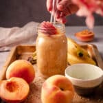 Peach smoothie in a mason jar on a tray with bananas and peaches.