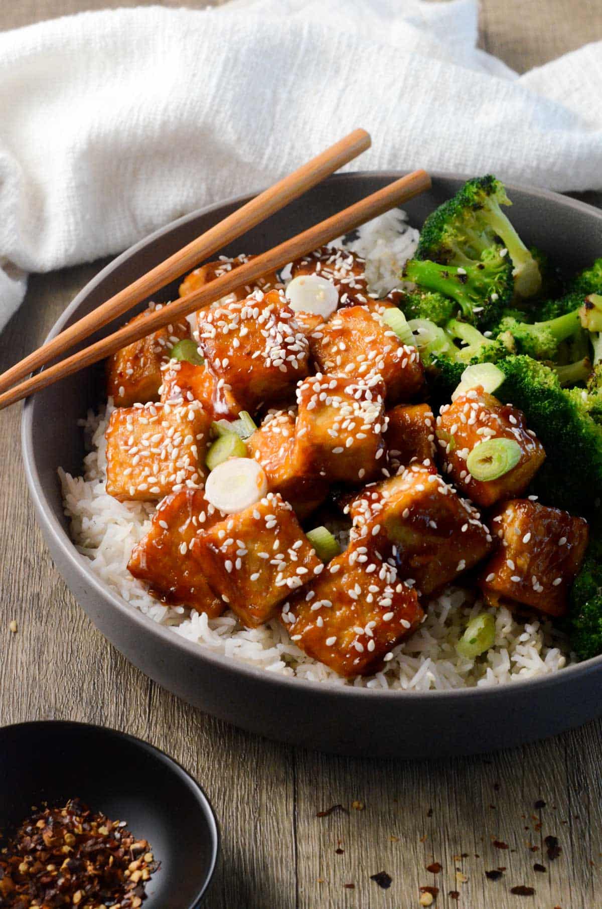 A bowl of tofu and broccoli with sesame seeds and chopsticks.