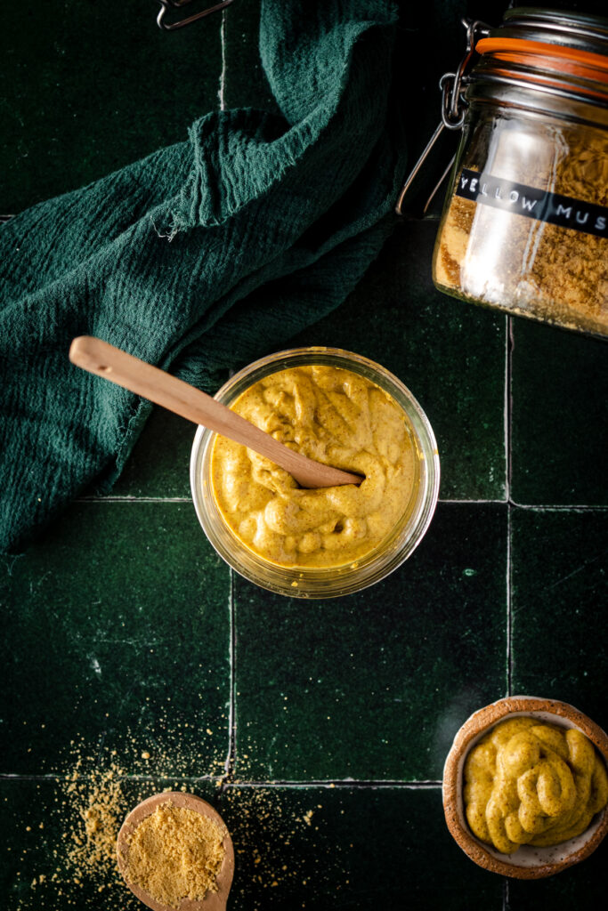 A jar of mustard with a spoon on a green tiled table.