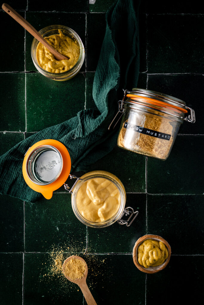 Two jars of mustard and a spoon on a tiled floor.