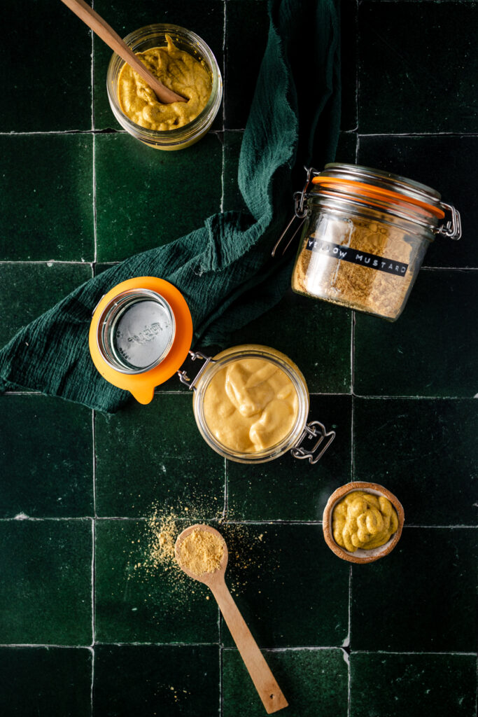 Two jars of mustard and a spoon on a green tile.