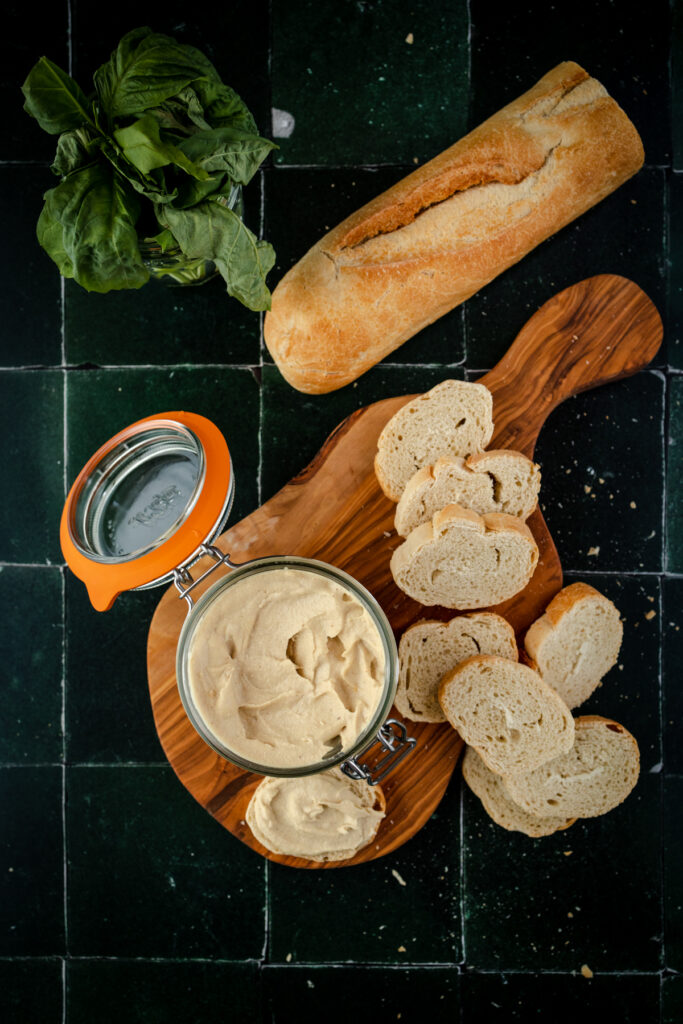 A wooden cutting board with bread and a jar of cashew ricotta.