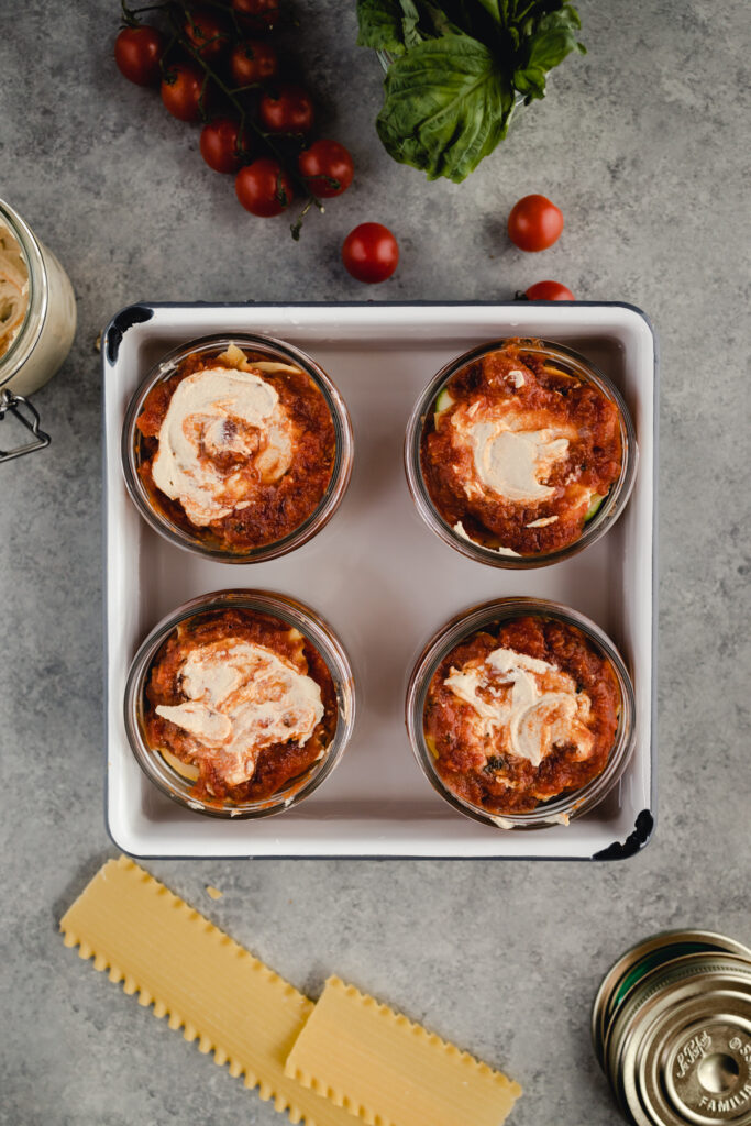 Prepared individual sized lasagnas in glass jars in a 9 by 9 pan with water.