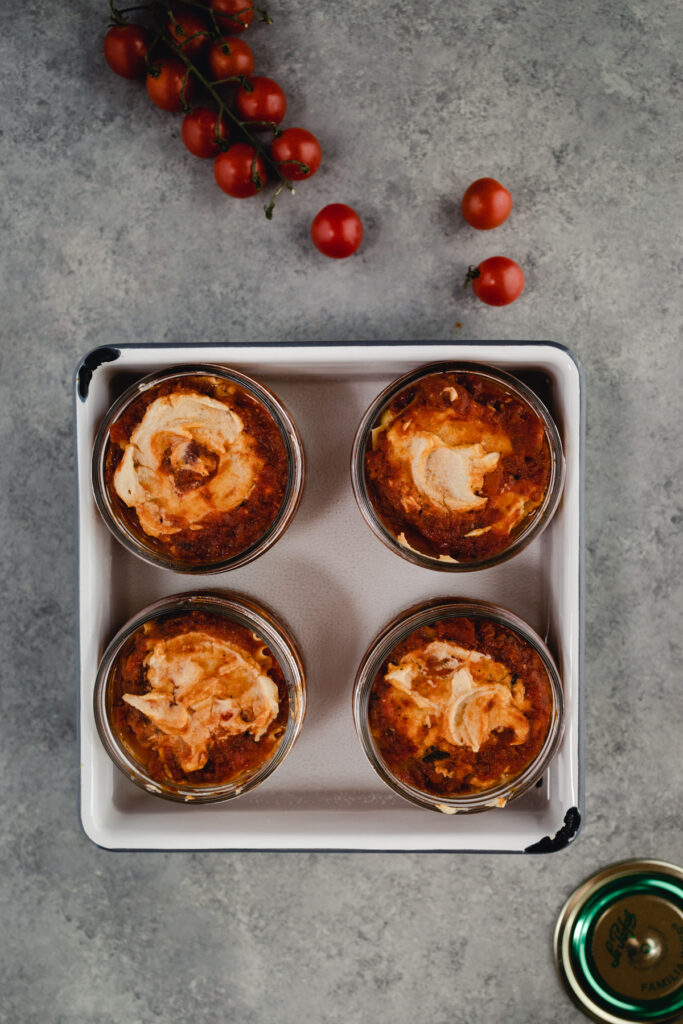 Baked personal sized lasagnas in glass jars in a 9 by 9 pan with water.