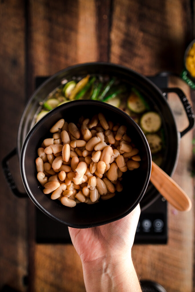 A hand holding a bowl of beans and vegetables on a wooden table.