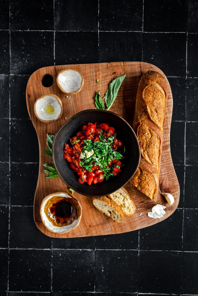 A bowl of tomatoes, bread and basil on a wooden cutting board.