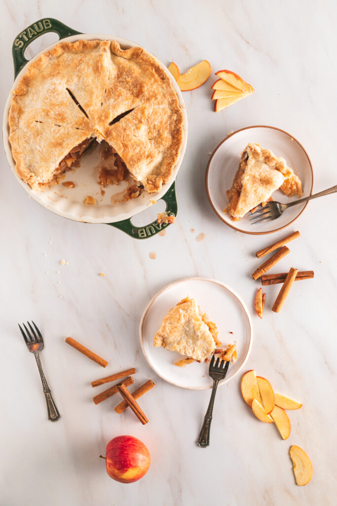 A slice of apple pie on a plate with cinnamon sticks.