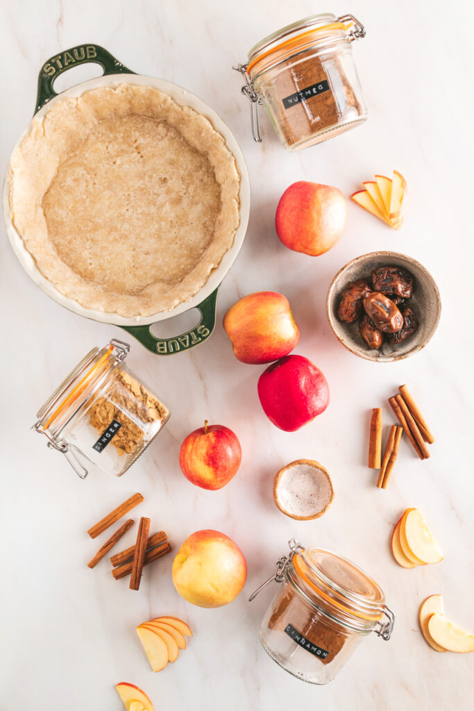 A pie crust, apples, and spices on a marble table.