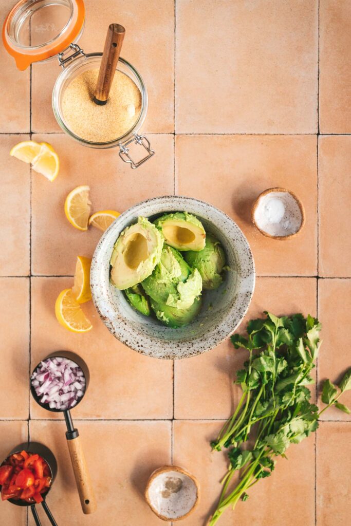 A bowl of avocado, onion, cilantro, and lime on a tiled floor.