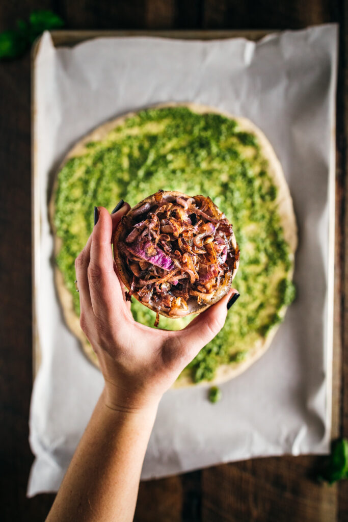 A person holding a pizza with a green pesto topping.