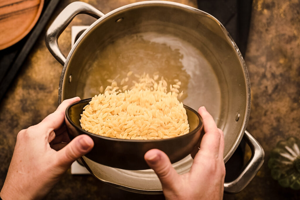 A person pouring orzo into a pan on top of a stove.