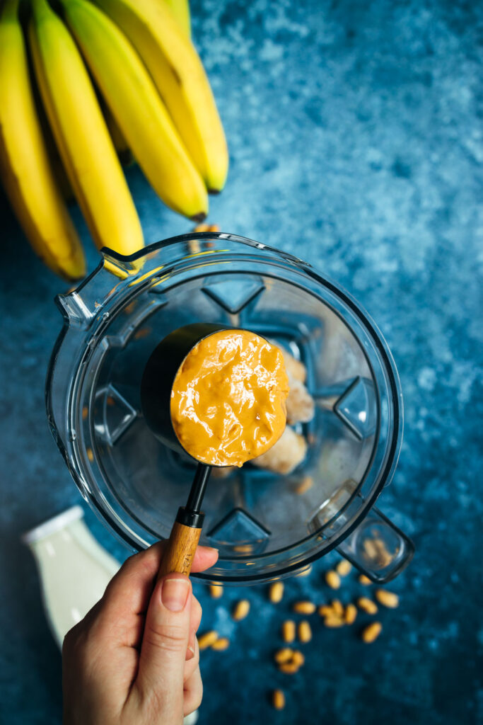 A hand is holding peanut butter in a blender with bananas in the background.