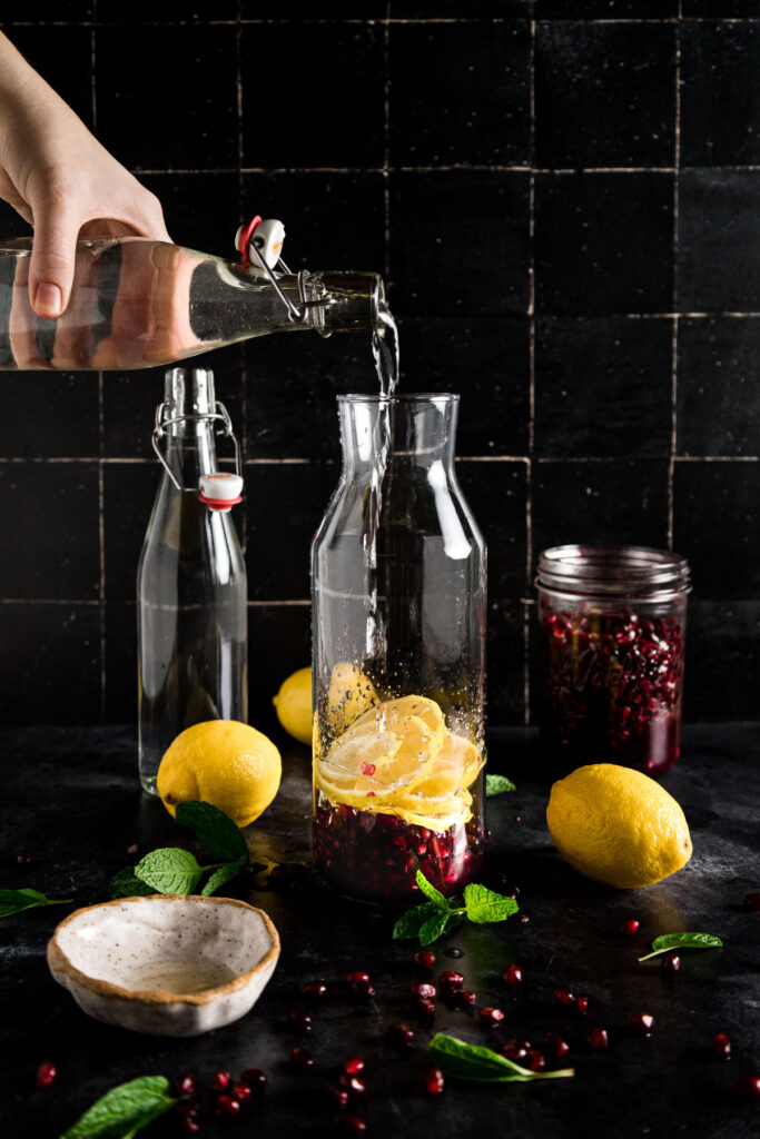 A person pouring water into a jar with lemons and pomegranate.