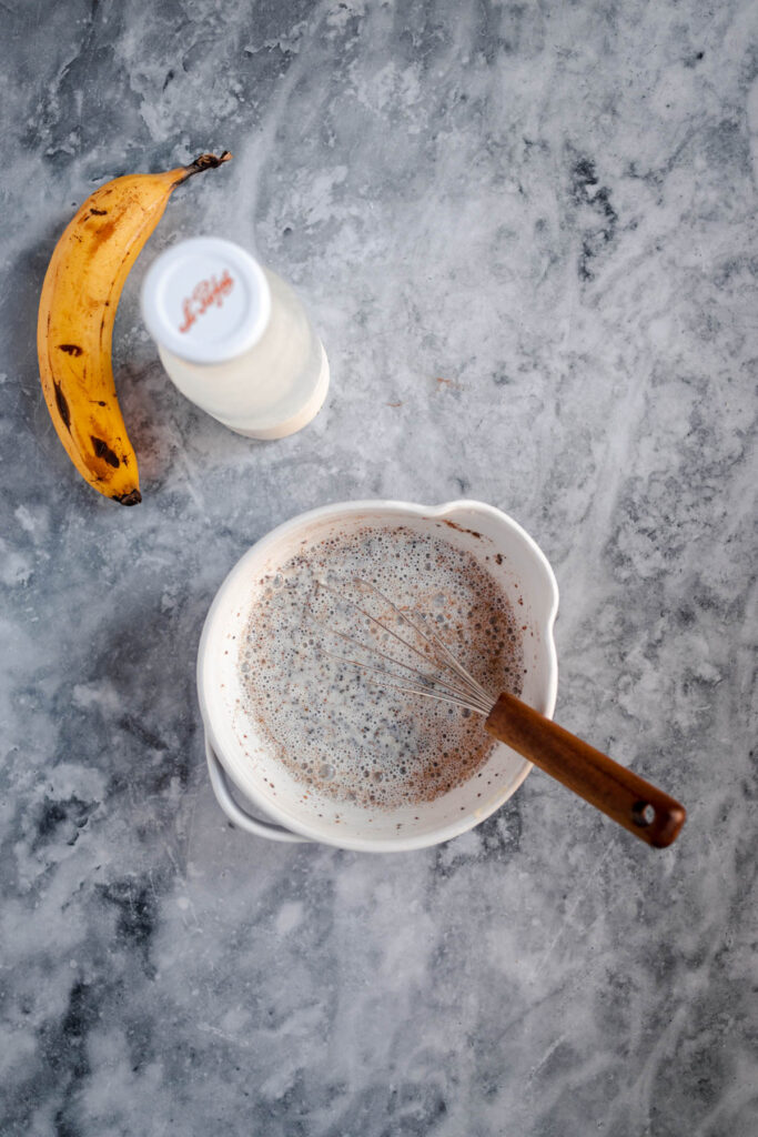 Bowl of batter with a whisk, a banana, and a bottle of milk on a marbled surface.