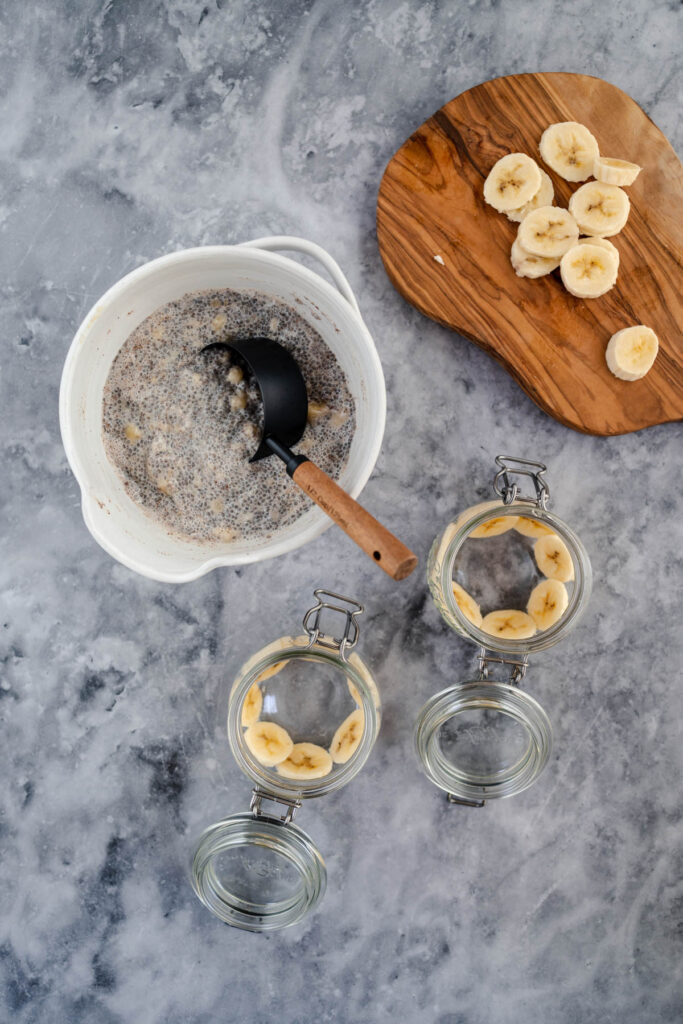 Overhead view of a bowl with oatmeal, a ladle, sliced bananas on a wooden board, and jars being layered with ingredients for a breakfast preparation.
