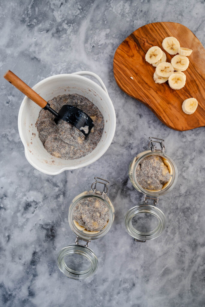 Ingredients for making banana chia pudding arranged on a marble countertop, with sliced bananas on a wooden board, a bowl of chia mixture, and empty glass jars ready for assembly.