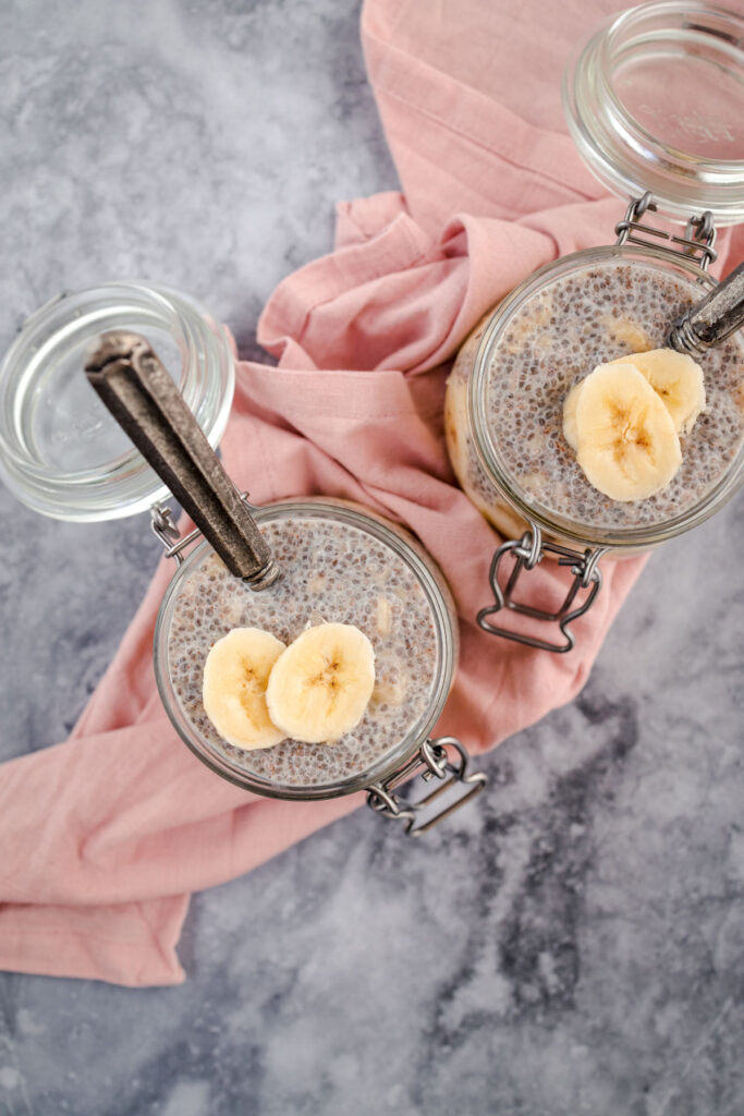 Two jars of chia pudding topped with banana slices and a cinnamon stick on a marble surface with a pink cloth.